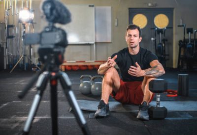 istockphoto-1176295972-612×612 Caucasian male fitness instructor sitting on exercise mat at gym and making vlog explaining how to workout with kettlebells and dumbbells.