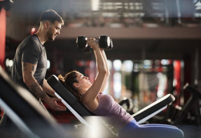 istockphoto-1392310150-612×612 Fitness instructor assisting athletic woman in exercising with dumbbells at gym.